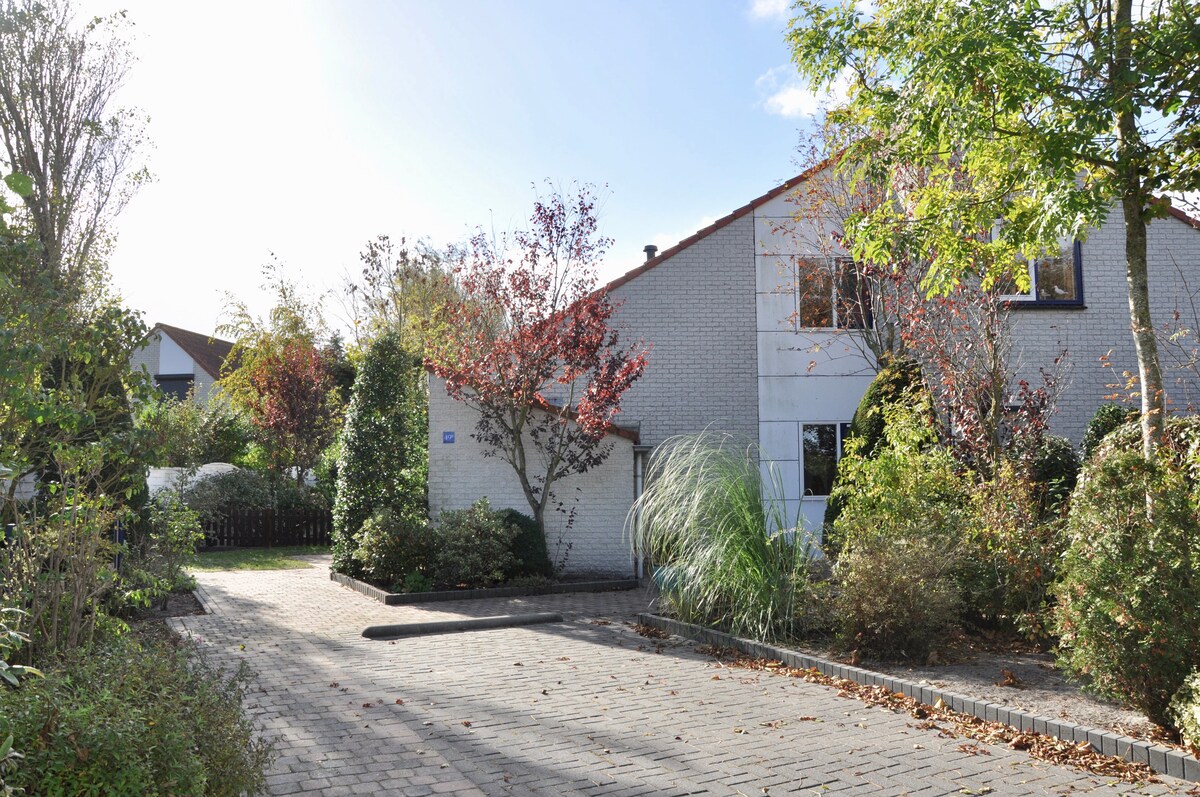 The exterior of the holiday home is depicted, featuring a light-colored façade with a well-kept garden. Surrounding trees and shrubs frame the entrance, while a paved driveway leads to the entrance. The clear sky and natural light contribute to an inviting atmosphere.