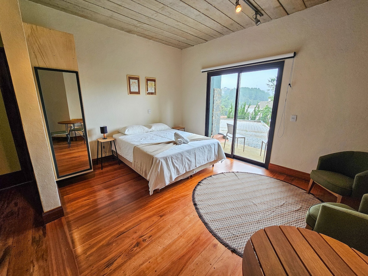 The spacious sleeping area features a large bed, complemented by a round woven area rug on polished wooden floors. A mirror is positioned against the wall, and a small table with chairs provides additional seating. Large sliding doors open to an outdoor view.