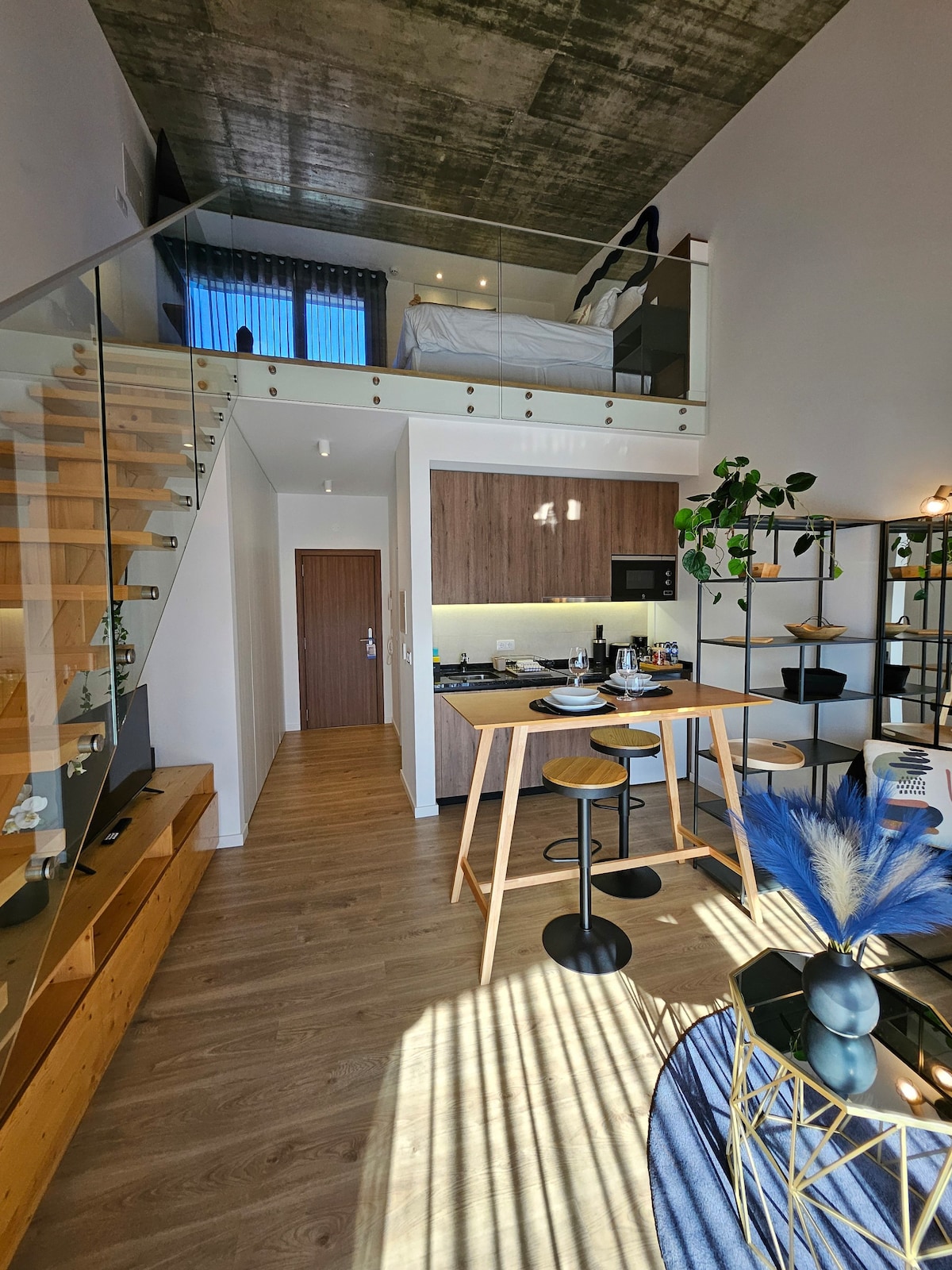 A bright loft apartment is depicted, showcasing an open living space with modern furnishings. The kitchen features a breakfast bar with stools, while a dining table is positioned nearby. A staircase leads to the upper bedroom area, which is illuminated by natural light from large windows.