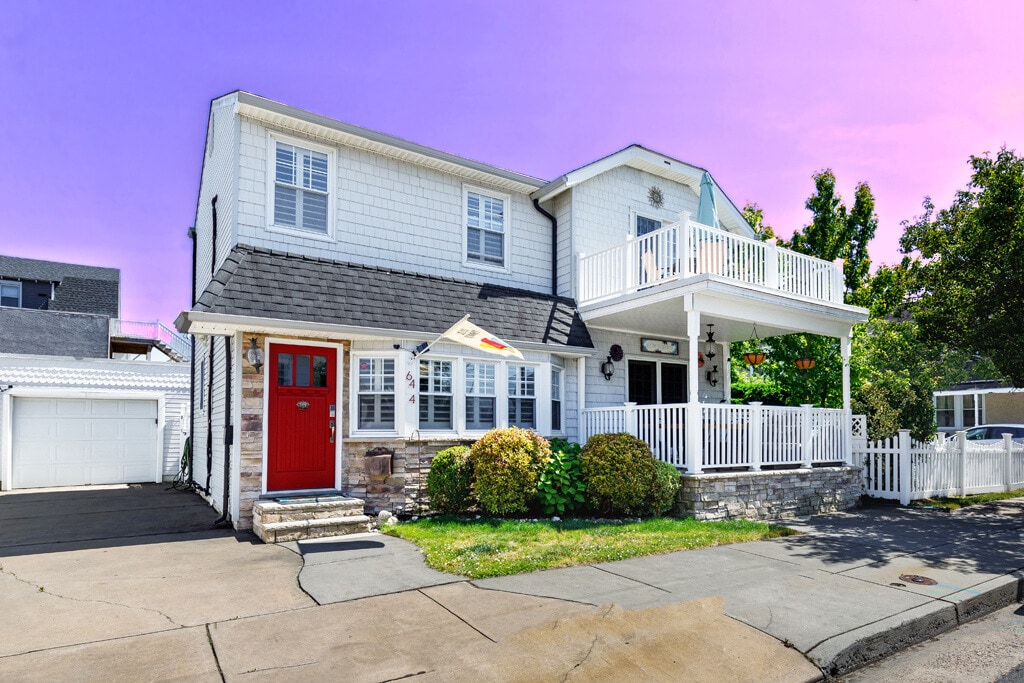 The exterior of a charming two-story home features white shingle siding and a red front door. A welcoming porch with railings provides an inviting entryway, while lush greenery and a well-manicured yard enhance the coastal appeal. The house is situated on a quiet street.
