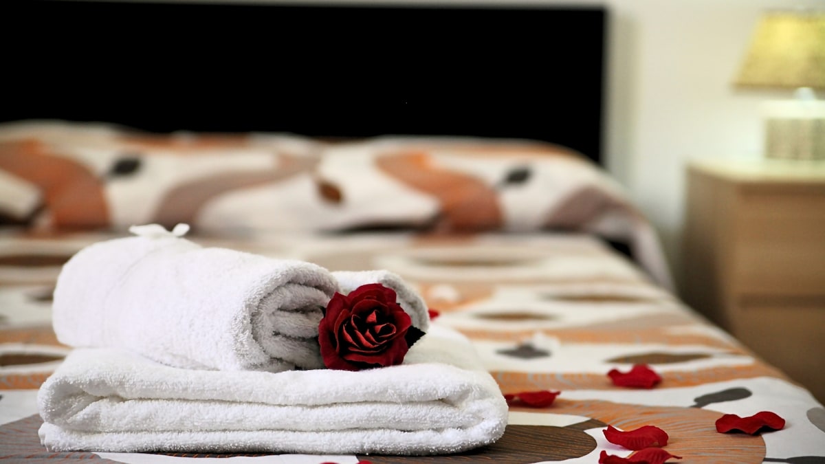 A neatly arranged set of towels is placed on a bed, complemented by a single rose and scattered petals. The bed features a patterned cover that adds a touch of character, with a dark headboard providing contrast. Soft lighting illuminates the bedside area.