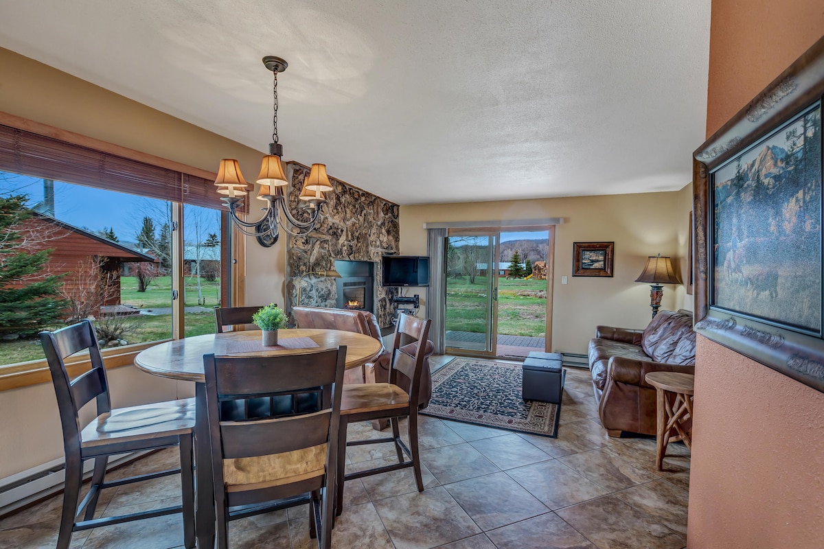 A dining area features a round table surrounded by wooden chairs, with a nearby window providing natural light. A comfortable seating arrangement is positioned beside a gas fireplace, and outdoor views of greenery and a playground are visible through the sliding glass doors.