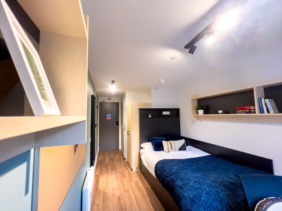 A compact bedroom features a small double bed with a navy blue blanket, complemented by light wooden shelving holding books and decor. Soft lighting illuminates the space, highlighting the smooth, wooden flooring. A door is visible at the end of the room, leading to the hallway.
