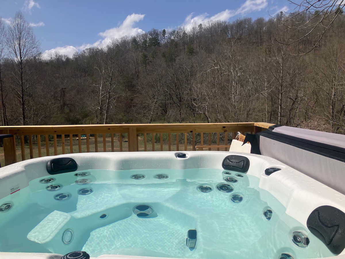 A four-seater hot tub is positioned on a spacious wooden deck, surrounded by a natural landscape of trees and hills. The clear water reflects the sky, and the area invites relaxation and enjoyment of the serene view.