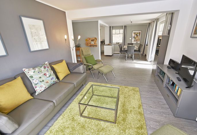 A spacious living area includes a comfortable sofa adorned with decorative cushions and a glass coffee table. A soft green rug covers the floor. An adjacent dining area is visible, featuring a table and chairs, with natural light filtering through the tall windows.