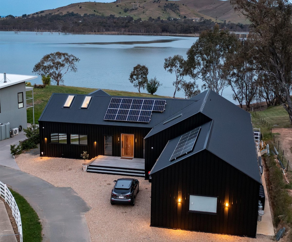 A modern home with a sleek black exterior is positioned near the calm waters of Lake Eildon. Solar panels line the roof, complementing the natural surroundings. A gravel driveway leads to the entrance, where warm lighting highlights the inviting facade.