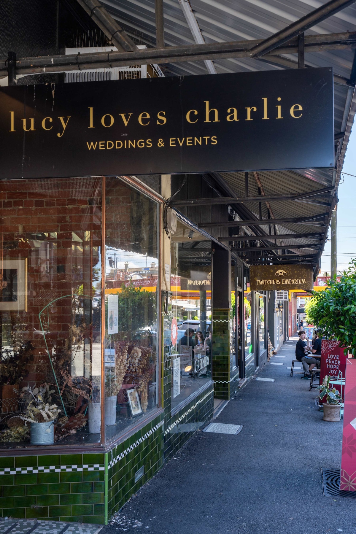 A charming streetscape features a row of boutique shops, including 'lucy loves charlie' with elegant signage. The storefront showcases well-arranged merchandise, and a pathway is lined with greenery, inviting visitors to explore the vibrant local scene.