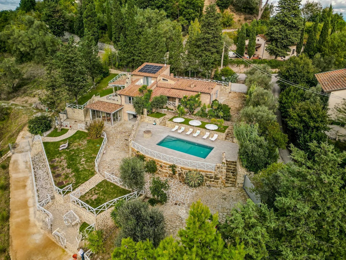 An aerial view showcases the independent villa nestled among lush greenery. A modern pool is surrounded by lounge chairs and stone pathways. The villa features a tile roof and terraces, integrating harmoniously with the natural landscape and offering a tranquil retreat.