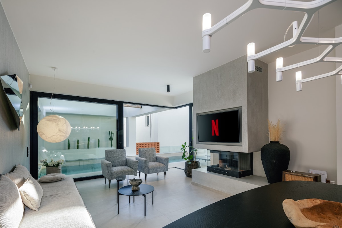 An open-plan living area features a minimalist design with a comfortable sofa and two armchairs, positioned near a large window that offers views of the private heated pool. A wall-mounted television is visible, with a sleek fireplace adding warmth to the ambiance.