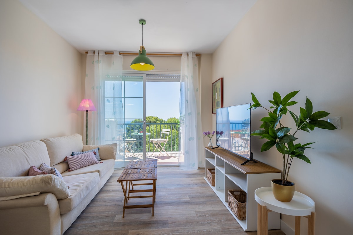 A light and airy living area is presented, featuring a soft beige sofa complemented by decorative cushions. A wooden coffee table rests on the floor, and a television is mounted on a white entertainment unit. Floor-to-ceiling glass doors lead to a sunlit balcony with outdoor seating.