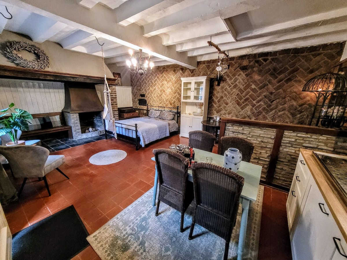 The open living area features a cozy sleeping space with a double bed, complemented by a textured wall design. A dining area with a round table and four woven chairs invites meals, while the kitchenette offers essential amenities. Natural light fills the space through multiple windows.