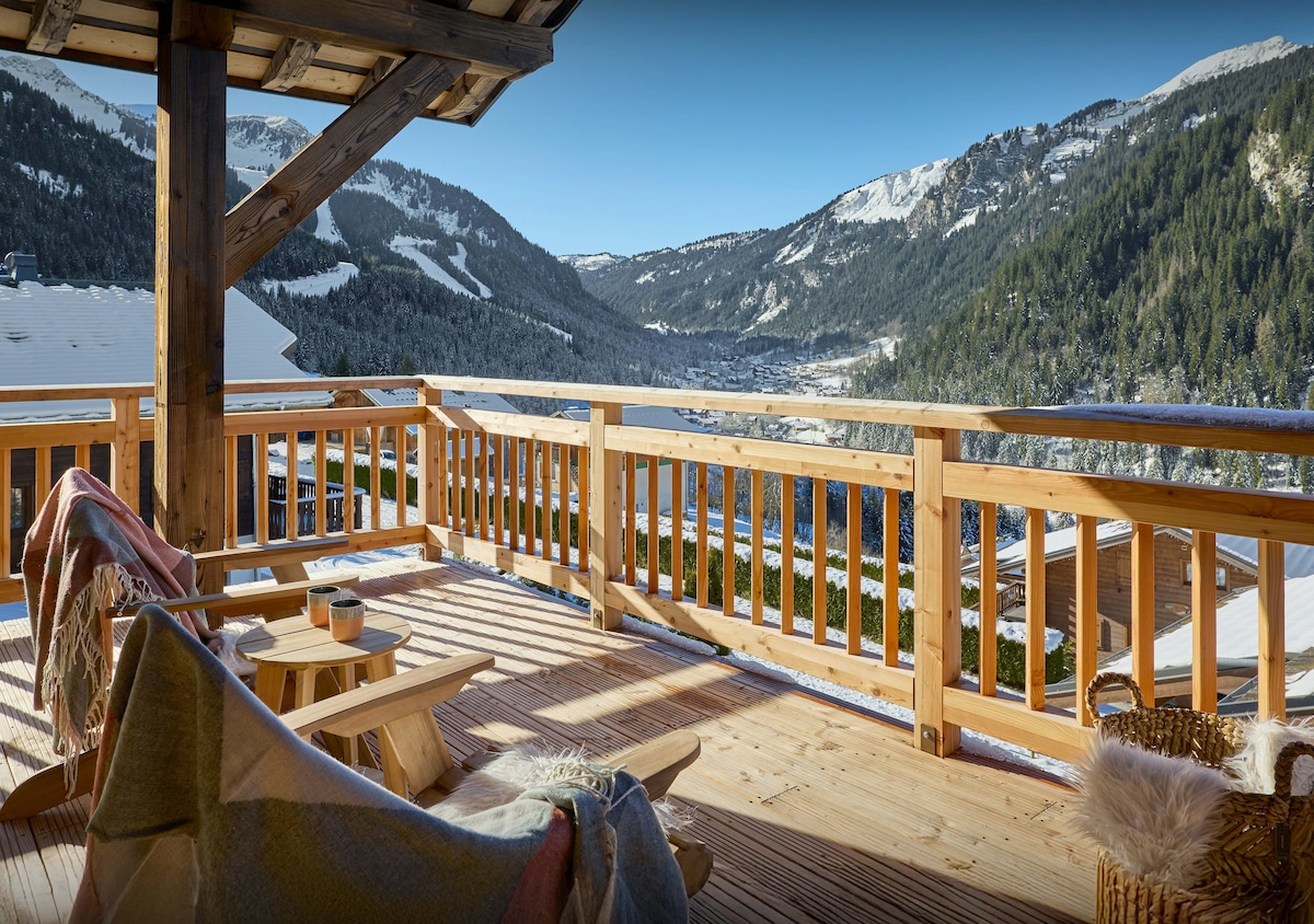 A wooden terrace is presented with expansive views of snow-covered mountains and valleys. Two casual chairs are complemented by a small round table, and colorful blankets are draped over the seating. Sunshine enhances the warm tones of the wooden railing.
