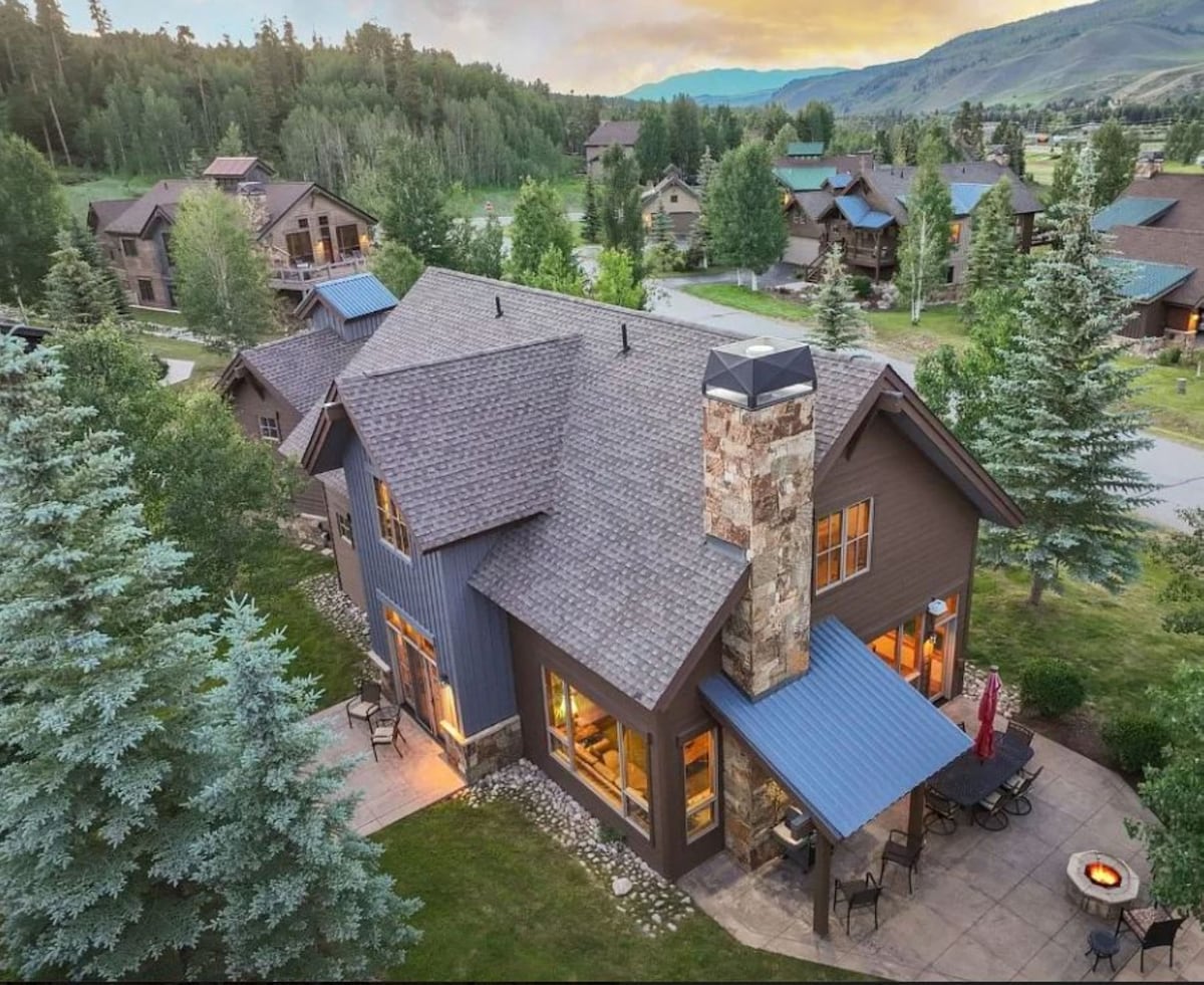 An aerial view captures the mountain retreat, showcasing a combination of wooden and stone materials. The sloped roofs blend with the natural surroundings, and a stone chimney stands prominently. Outdoor seating is visible on the patio, surrounded by lush greenery and scenic hills.