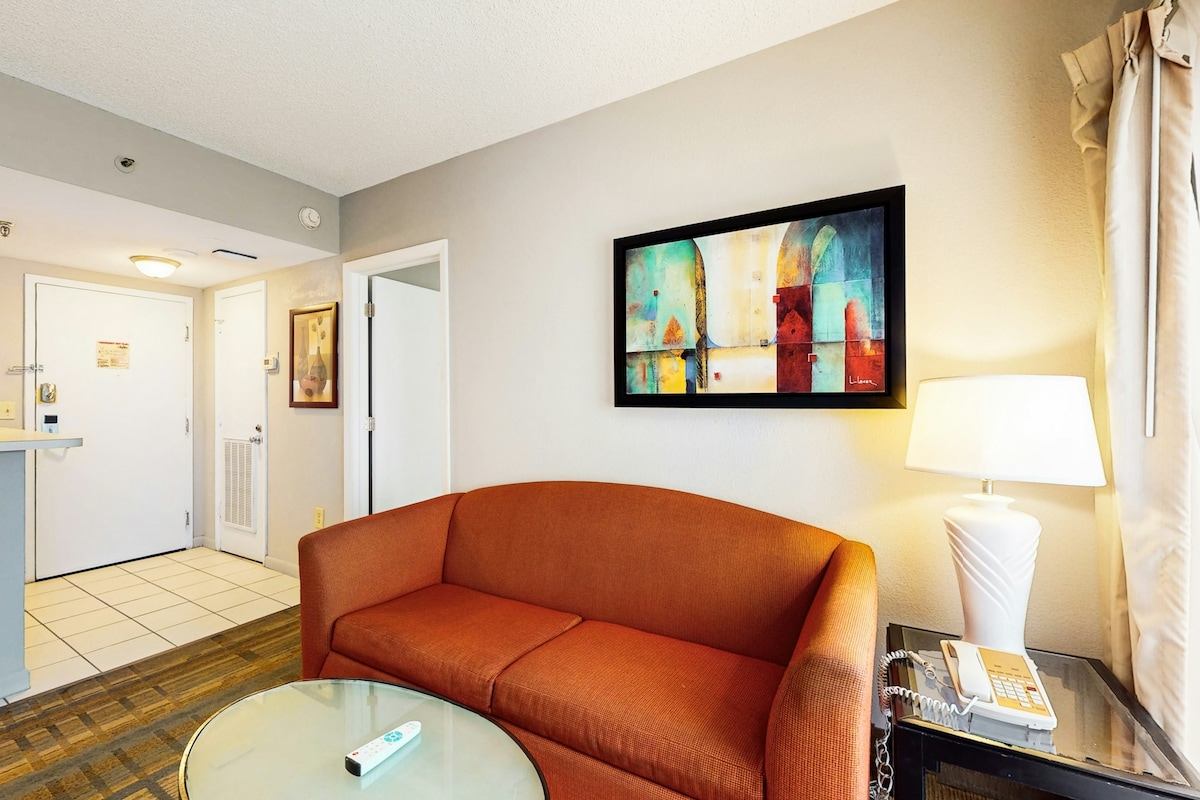 A cozy living area features an orange couch complemented by a glass coffee table. A colorful artwork hangs above the couch, while soft lighting is provided by a nearby lamp. The entrance to the condo and a kitchenette are visible in the background.