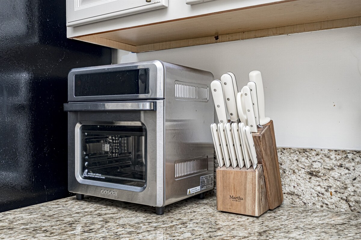 A modern stainless steel air fryer stands next to a wooden knife block, which holds a complete set of white-handled knives. The countertop features a textured granite surface, creating a clean and organized kitchen space.