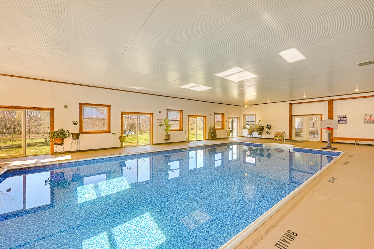 A spacious indoor pool area features a large, heated pool with shimmering water reflecting natural light. Surrounding windows provide views of the outdoors, and several potted plants add a touch of greenery to the light-filled space.