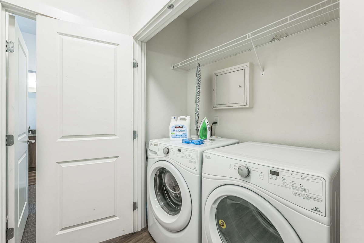 A laundry area is equipped with a washing machine and a dryer, each positioned beside a shelf for laundry supplies. The room features light-colored walls and has a door leading to another area, contributing to the functional layout of the unit.