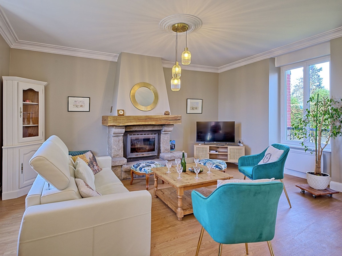 The living room showcases a modern design with a stone fireplace as a focal point. Comfortable seating includes a white sofa and teal accent chairs. A flat-screen TV is positioned on a wooden unit, and natural light floods the space through large windows.