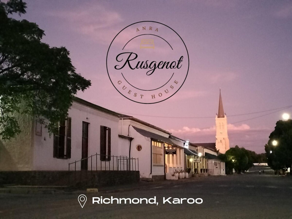 The image depicts a street view of a guest house, featuring a row of single-story buildings under a violet sky at dusk. A steeple of a church is visible in the background, with trees lining the street creating a shaded ambiance.
