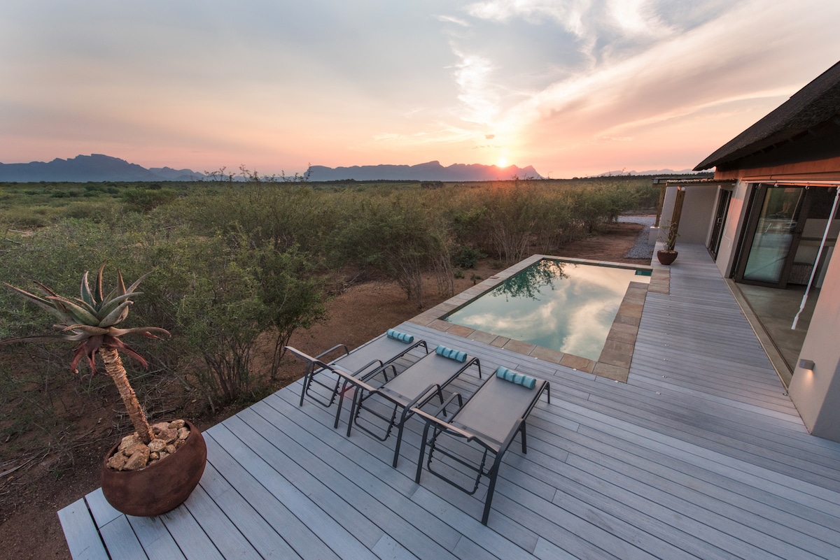 A spacious wooden deck provides access to a heated swimming pool, with sun loungers placed nearby for relaxation. The scene captures a stunning sunset over the Little Drakensberg mountains, framed by lush vegetation, while the villa structure stands elegantly in the background.