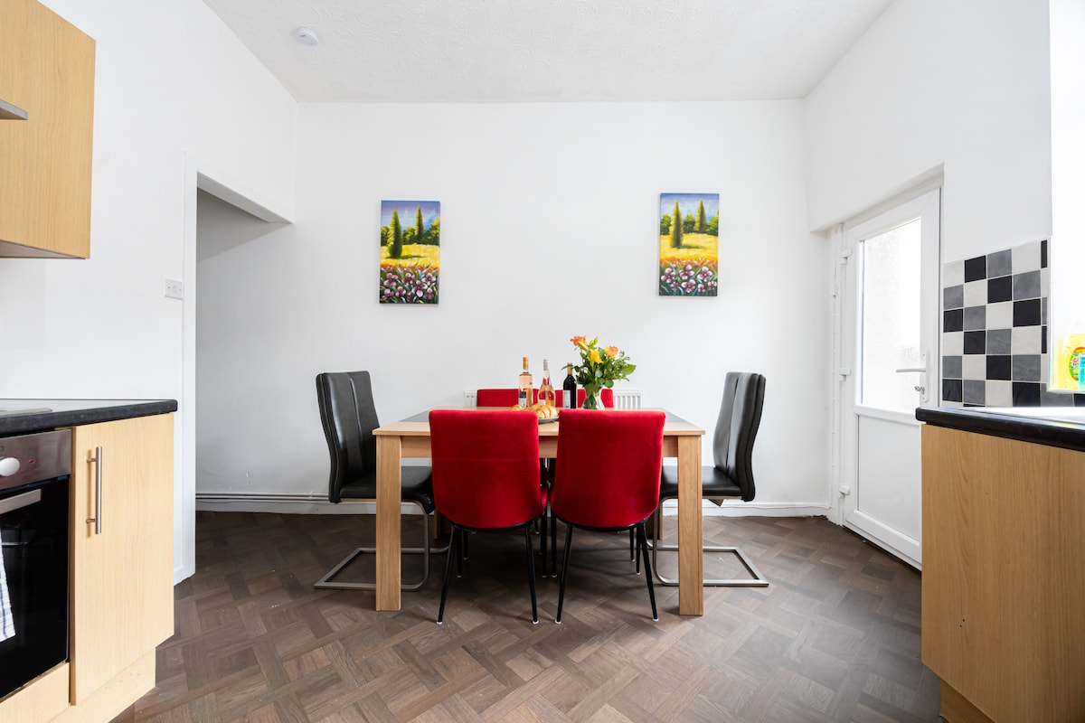 A well-lit dining area features a wooden table surrounded by six chairs in a mix of red and black. Two vibrant landscape paintings adorn the walls, and a bouquet of flowers sits in the center, adding a fresh element to the space.