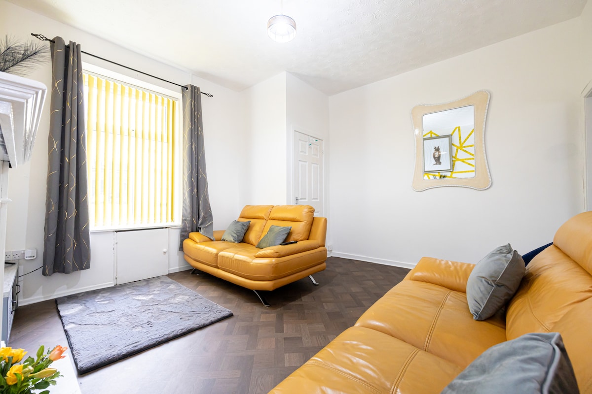 A bright living room is showcased with large windows allowing ample natural light. A yellow leather sofa is positioned next to a coordinating armchair, complemented by a plush gray rug. A mirror with a unique design hangs on the wall, adding depth to the space.