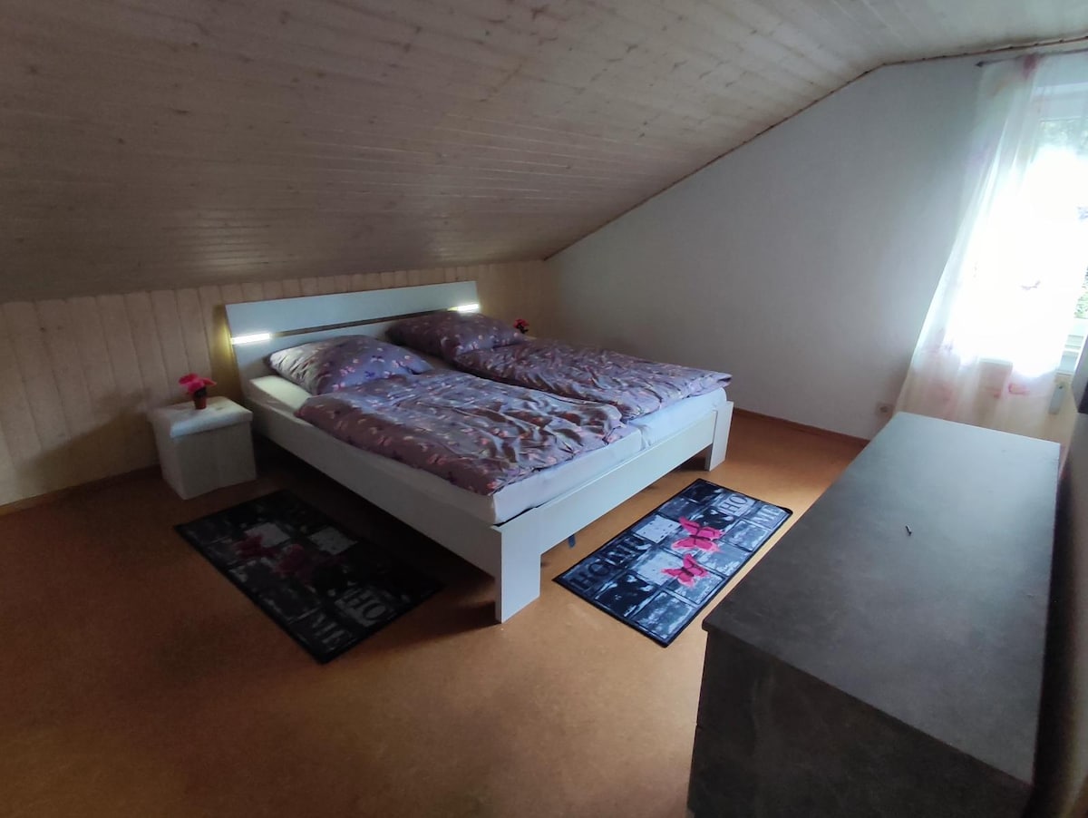 The sleeping area features a spacious bed with soft bedding and two decorative rugs beneath it. A bedside table holds a small flower vase, while natural light enters through a nearby window adorned with sheer curtains. A simple dresser adds functionality to the room.