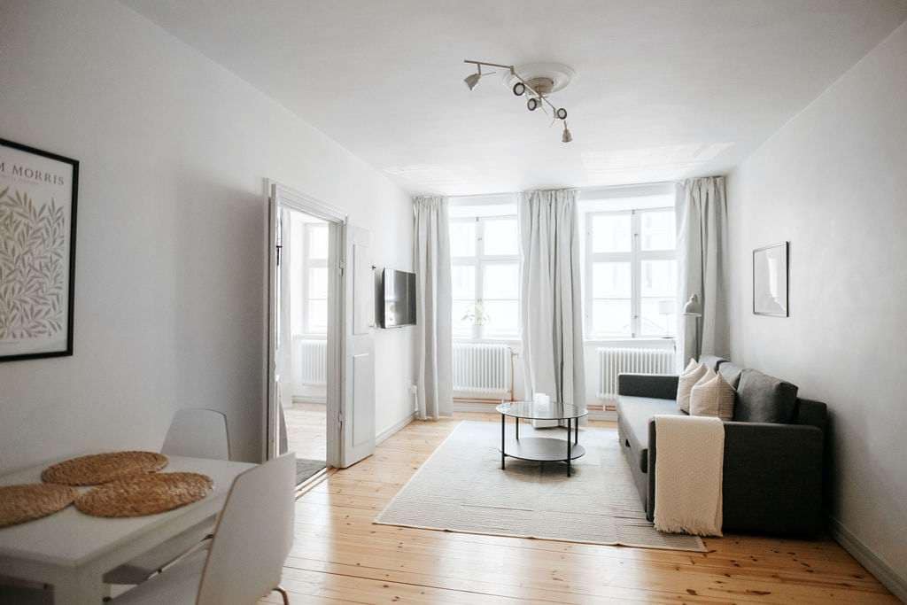 A bright living area features light walls and wooden floors, accented by a gray sofa and circular coffee table. Natural light filters through large windows, dressed with light curtains. A dining table set for four complements the space, along with wall art adding visual interest.