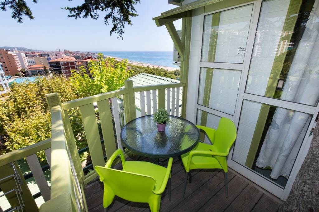 An outdoor terrace is furnished with a glass-top table and two bright green chairs. A small potted plant is centered on the table. The view features the coastline and ocean in the distance, framed by trees and nearby buildings.