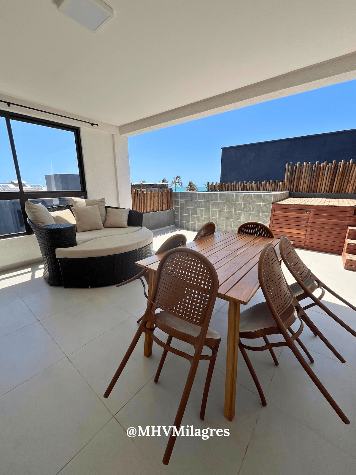 An outdoor dining area features a wooden table surrounded by six woven chairs. A lounge seating area with a circular sofa complements the space, offering a comfortable spot to enjoy views of the surrounding area. Natural light brightens the contemporary design.