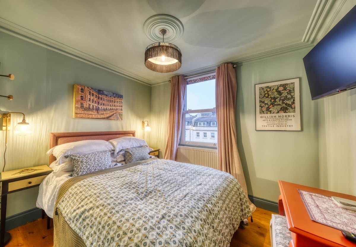 A cozy double room features a spacious bed covered in a patterned quilt. Soft green walls complement decorative artwork. Natural light enters through a large window, and a TV is mounted on the wall. A bedside table with a reading light is positioned on one side.