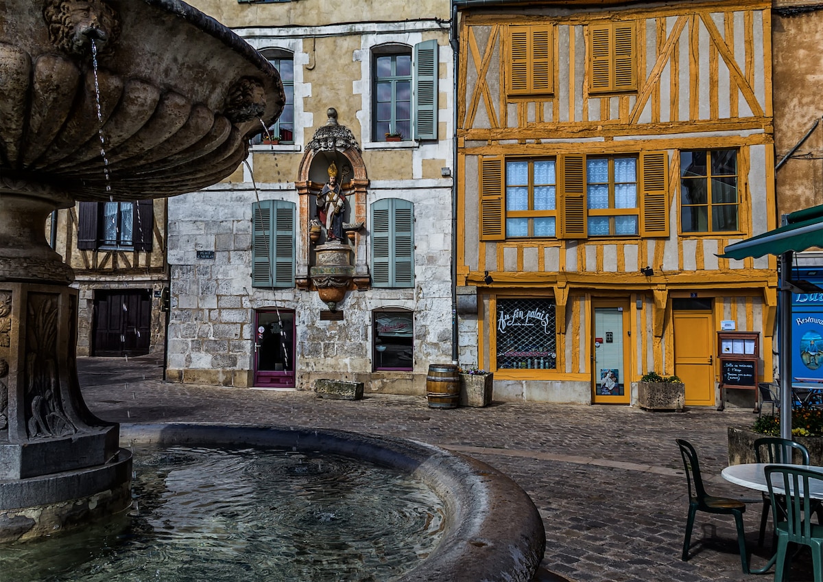 A historic square features a stone fountain in the foreground, with a statue atop it. Surrounding buildings display traditional architecture, including wooden beams and colorful shutters. The atmosphere is quietly vibrant, with cobblestone paths leading through the area.