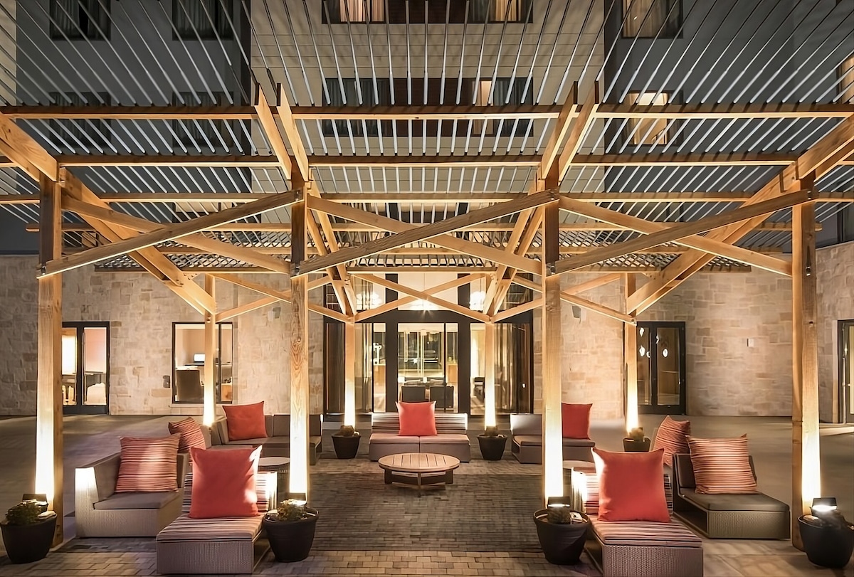 An outdoor lounge area is framed by a wooden pergola, featuring cozy seating with soft red cushions. Ambient lighting creates a warm atmosphere, while a circular coffee table sits at the center. Large glass doors in the background provide access to the interior.