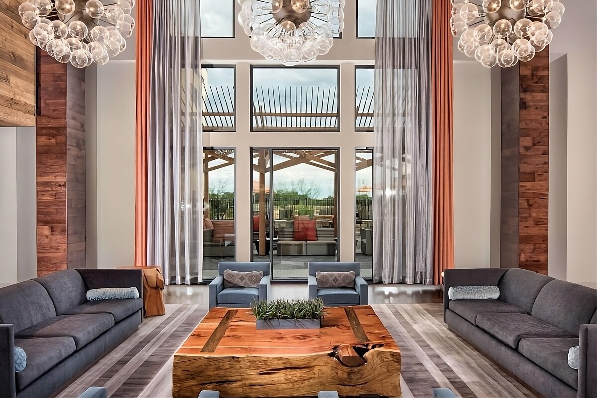 The lobby area showcases a modern design featuring large windows that allow natural light to fill the space. Two plush gray sofas face a central wooden coffee table, adorned with greenery. Elegant light fixtures hang from the ceiling, complementing the warm tones of the decor.