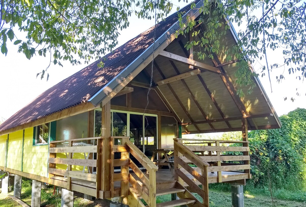 The bungalow features a sloped roof with a wooden terrace, surrounded by lush greenery. Large windows provide views of the garden while allowing natural light into the living space. The entrance is accessible via wooden steps leading to the inviting outdoor area.