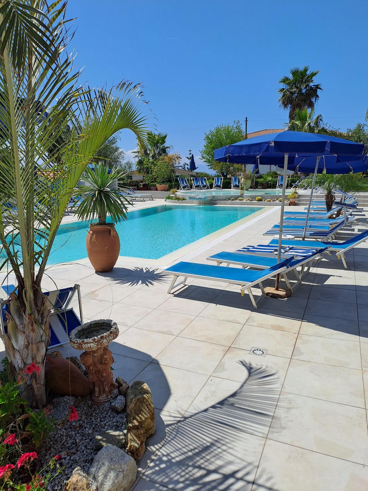A shared outdoor area features a large pool surrounded by sun loungers and blue umbrellas. Tall palm trees and colorful plants add to the landscape. A decorative fountain is visible near the pool, enhancing the tranquil atmosphere of the space.