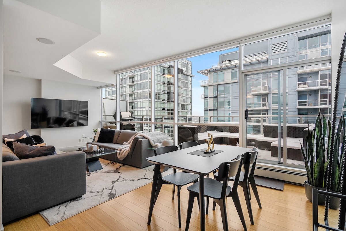 The living area is filled with natural light from large windows that overlook the city. A modern dark sofa and coffee table create a relaxed seating space, while a dining table accompanies stylish black chairs. A sleek TV is mounted on the wall, enhancing the contemporary design.