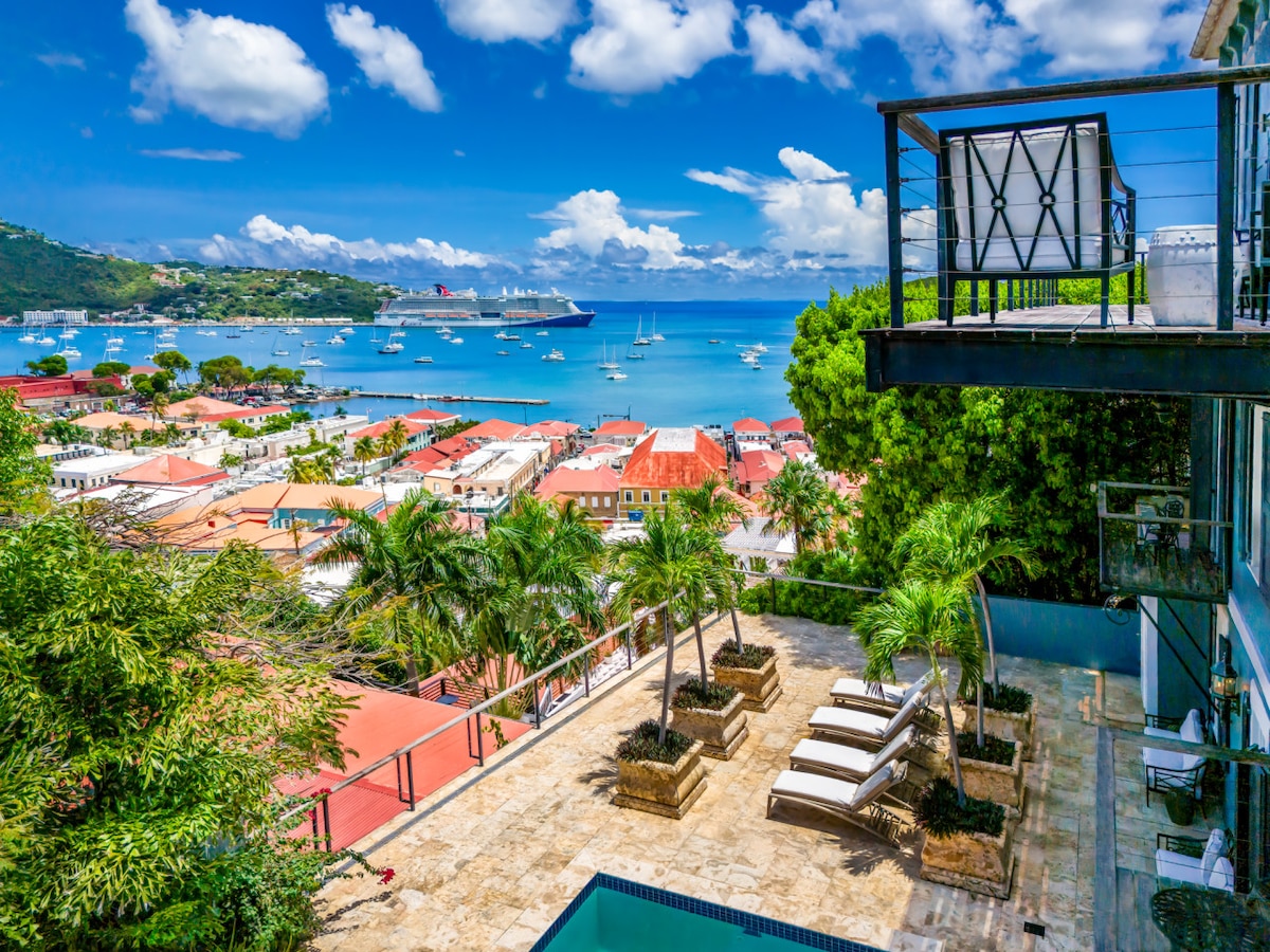 A sweeping view captures the vibrant Charlotte Amalie harbor, with boats anchored in turquoise waters. Lush palm trees frame the image, and the terrace features lounge chairs beside a tranquil pool, inviting relaxation while enjoying the scenic backdrop of colorful rooftops and distant hills.