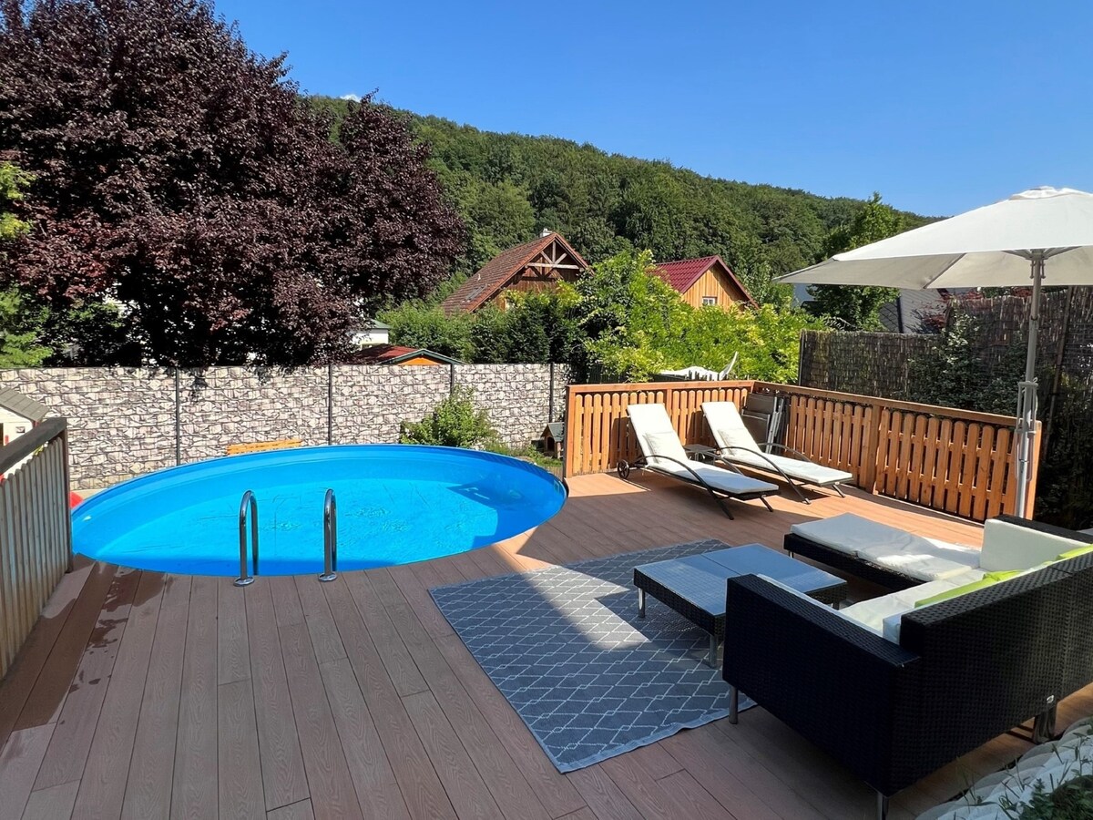 An outdoor area features a private, round swimming pool surrounded by a wooden deck. Two lounge chairs are positioned beside the pool. A shaded seating area with a modern lounge set is also visible, along with an umbrella providing protection from the sun.
