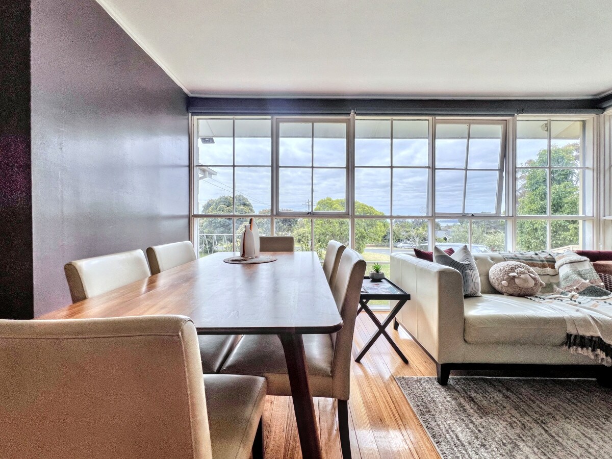 An open-plan living and dining area captures natural light through large windows, framing a view of lush greenery. A wooden dining table is surrounded by beige chairs, while a soft white sofa adds comfort to the space, complemented by decorative pillows and a textured throw.