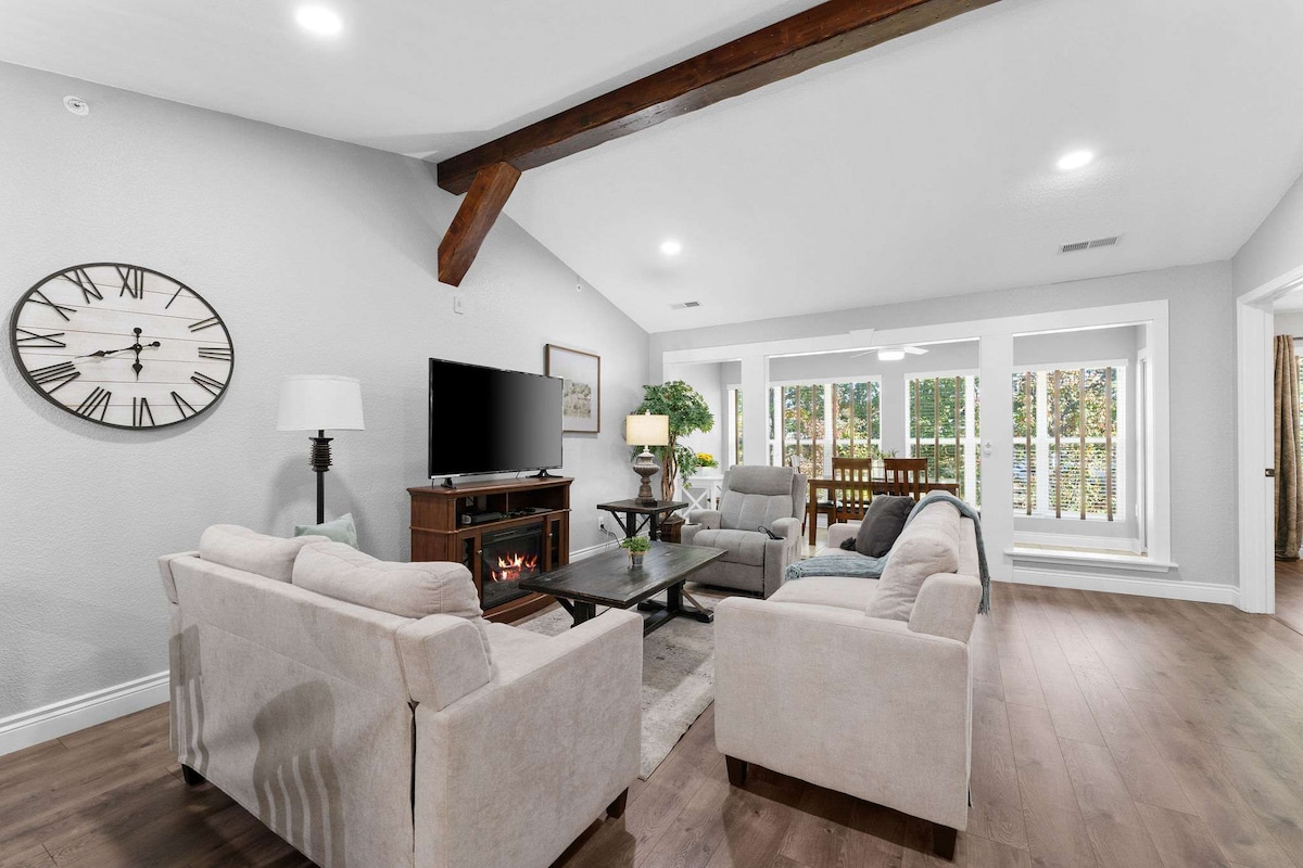 The living room features comfortable seating with light-colored sofas facing a coffee table. A television is mounted on a wall unit. Large windows allow natural light to fill the space, which is accented by warm wood beams overhead and decorative plants in the corners.