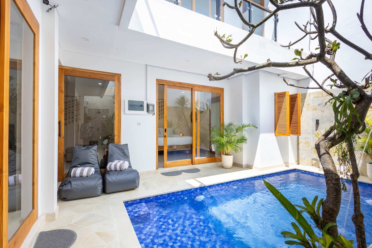 A private outdoor area showcases a refreshing pool surrounded by natural stone and tropical plants. Large glass doors open to an airy interior space, while comfortable bean bags are positioned nearby for relaxation. Sunlight reflects off the water, creating a serene atmosphere.