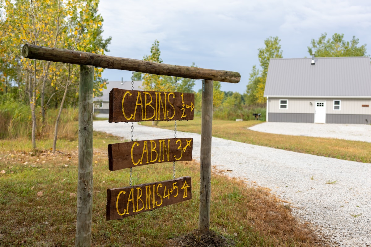 A wooden signpost directs guests towards the cabins, with clearly marked arrows indicating the path to Cabin 1, Cabin 3, and Cabins 4 and 5. The background features a well-maintained gravel road surrounded by greenery and a cabin with a modern metal roof.