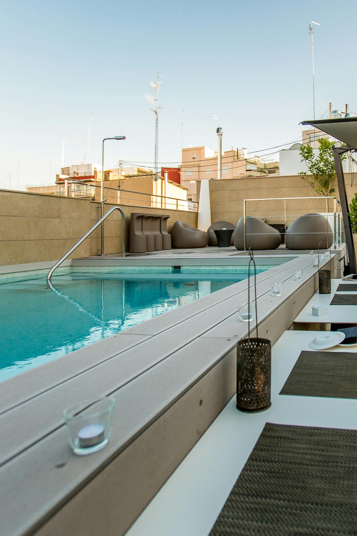 A rooftop area features a modern plunge pool surrounded by elegant lounge seating. The pool is framed by a wooden deck, with plush chairs providing a comfortable setting for relaxation. A lantern and decorative accents enhance the tranquil ambiance, complemented by a clear sky above.