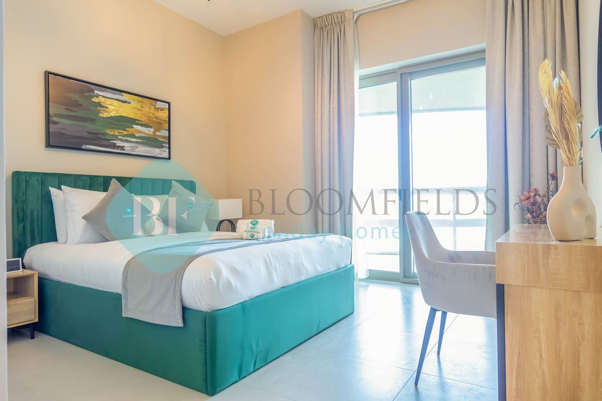 A spacious bedroom features a comfortable bed dressed in crisp linens, with decorative pillows. Neutral tones create a calming environment. A wooden desk is positioned near a large window, which offers natural light and views of the outside. Soft curtains frame the window.
