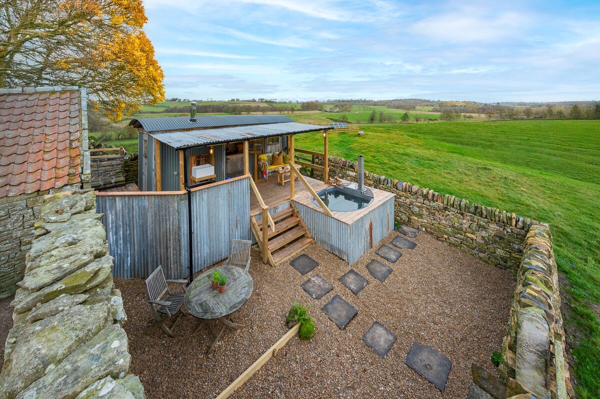 The rustic cabin is surrounded by expansive green fields, featuring wooden steps leading to a welcoming verandah. A wood-fired hot tub is positioned nearby, complemented by a circular stone table and chairs set on gravel. The serene landscape promotes a tranquil retreat.