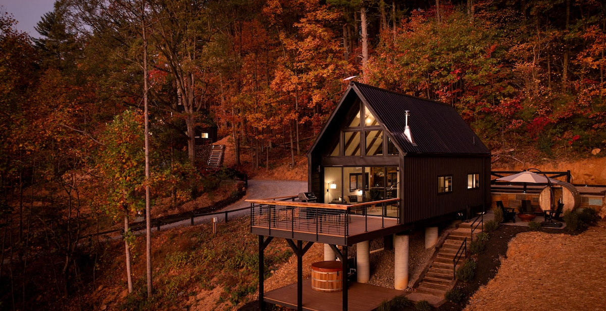 The cabin's exterior displays a modern A-frame design complemented by warm autumn foliage. A multi-level deck features a hot tub, while large windows provide views into the inviting living space. The driveway is visible, winding through the surrounding natural landscape.