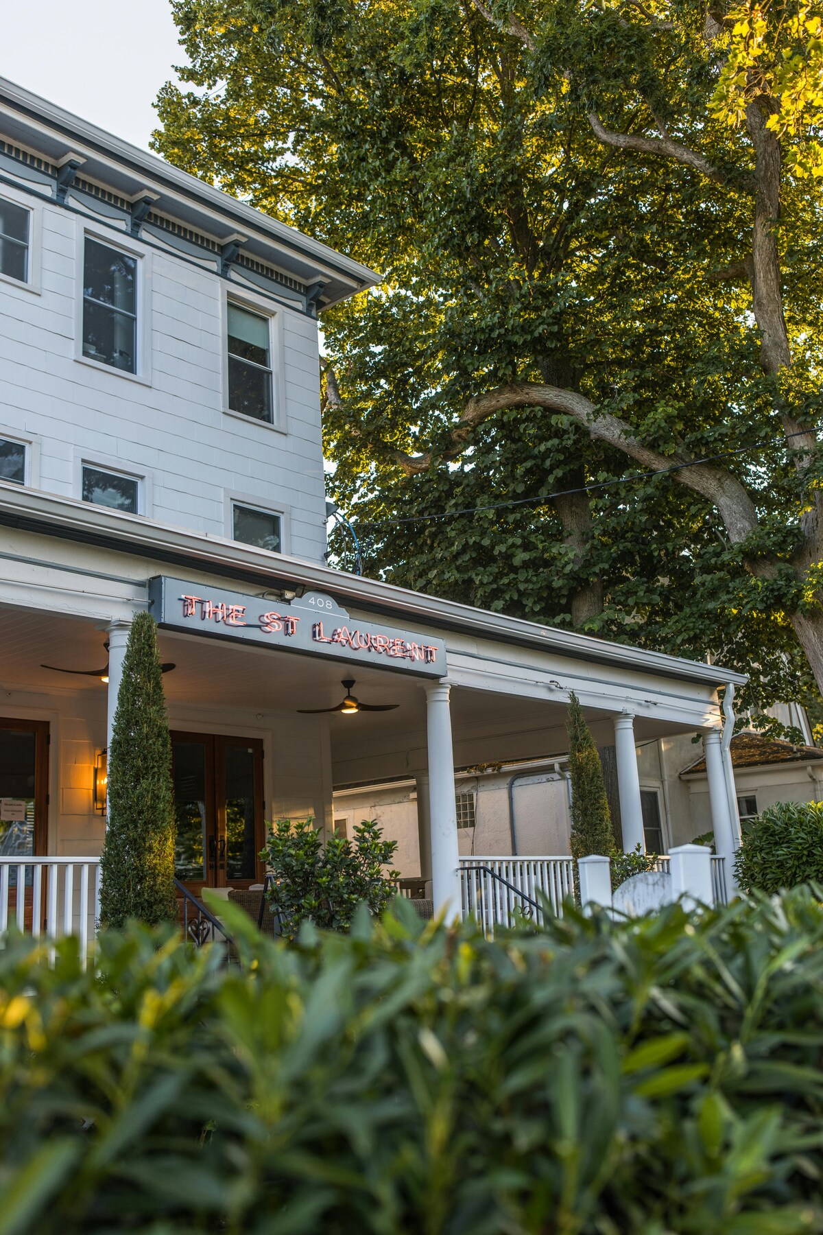 The exterior of The St. Laurent is shown, featuring a classic structure with a welcoming porch. Lush greenery surrounds the entrance, and tall trees provide shade. The name 'The St. Laurent' is prominently displayed above the doorway, enhancing the inviting atmosphere.