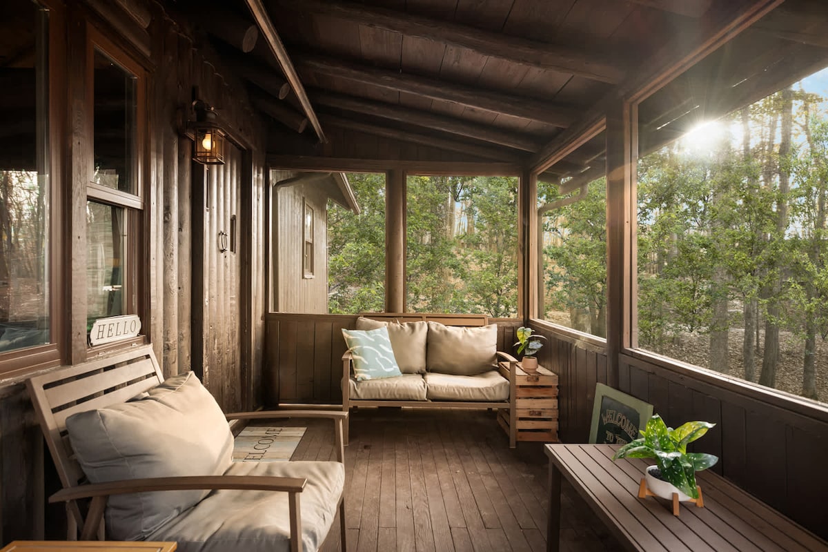 A screened-in porch is shown with wooden walls and flooring, offering a cozy seating area. Cushioned sofas are complemented by a small wooden table and a potted plant. Natural light filters through the surrounding trees, providing a serene environment.