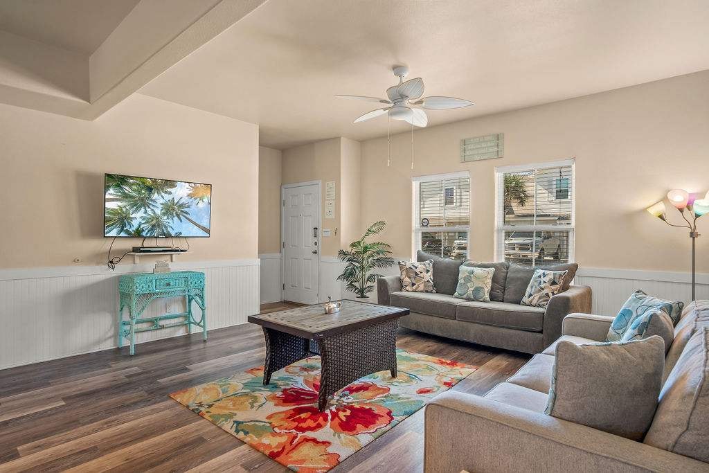 The living room features a comfortable seating area that includes a large gray sofa and a smaller loveseat, positioned around a coffee table. A flat-screen TV is mounted on the wall, with natural light streaming in through the windows. A vibrant area rug adds color to the space.